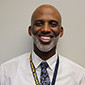 A man smiles. He has a close cropped beard and is wearing a dress shirt and tie.