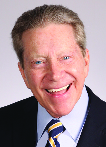 A man with light hair, wearing a blue and gold striped tie, blue shirt and dark suit jacket. He is smiling broadly and has blue eyes.