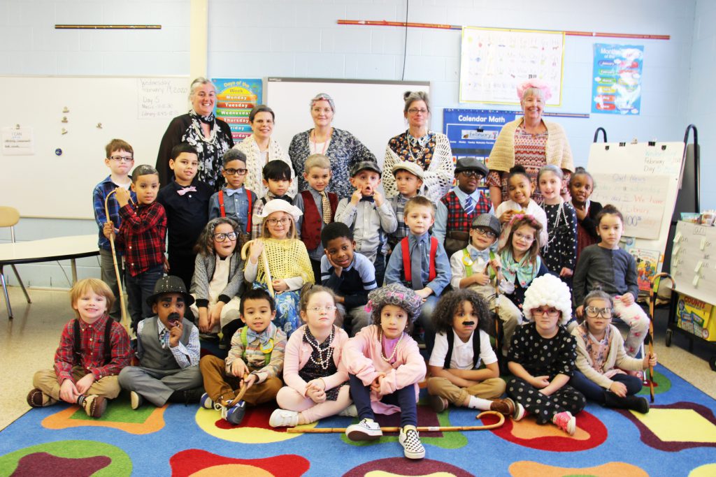 A class of 27 kindergarten children sit and stand in three rows. Many are dressed as 100-year-old people. Five adults stand behind them.