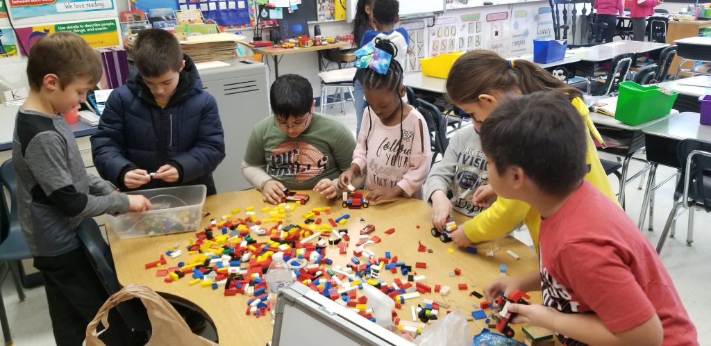 A group of five elementary students sit and stand around a table filled with Legos as they build things