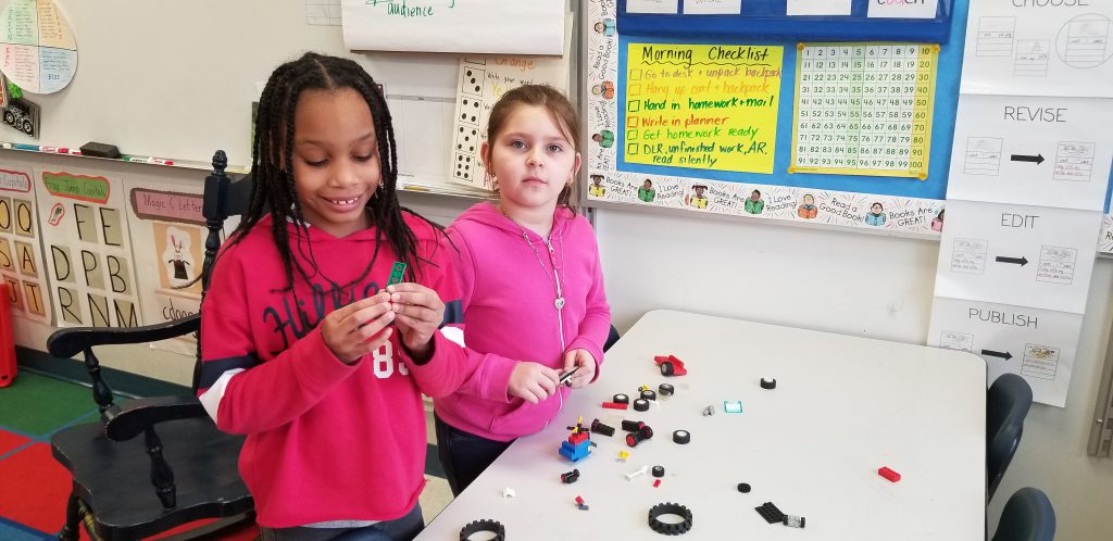 Two elementary girls, one wearing red and the other pink, build with Legos.