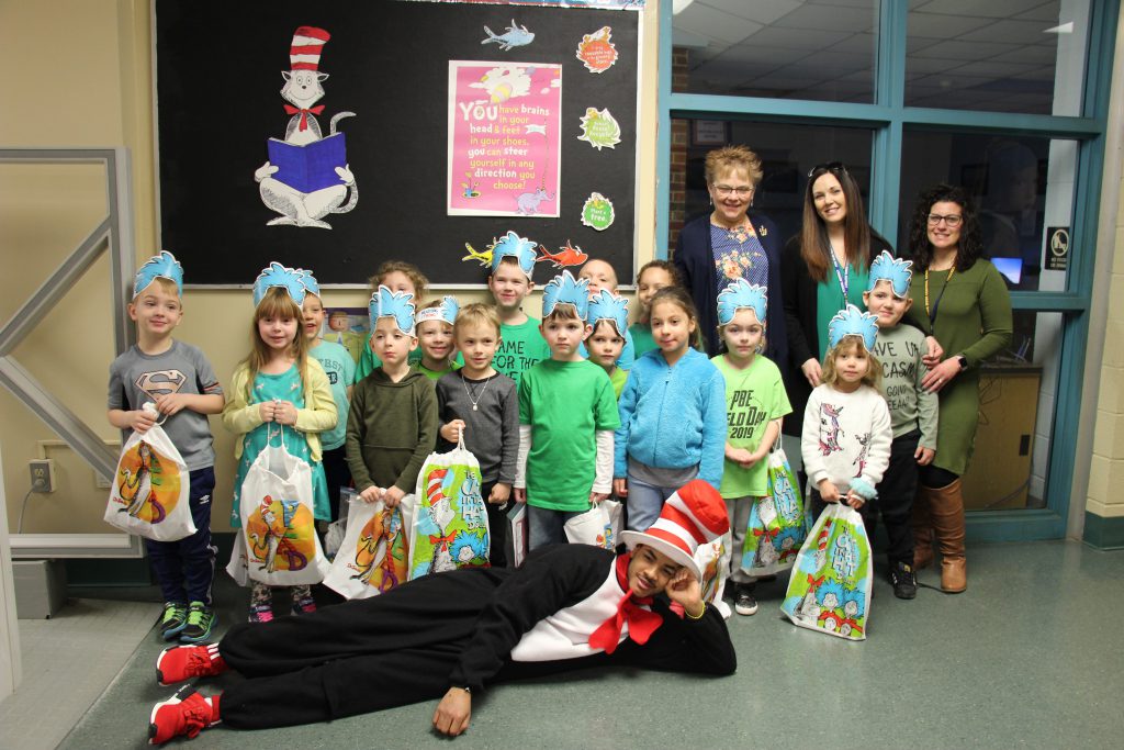 A kindergarten class wearing paper hats stand and smile as a high school student dressed as the cat in the hat lays in front of them.