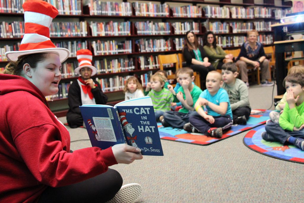 A high school girls sitting on the floor wearing a red and white striped hat, holding the Cat in the Hat book up while a large group of kindergarten students sit listening on the floor.