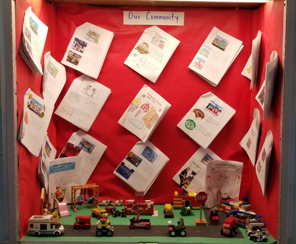 A display case with a red background has a lego city on the bottom and more than a dozen papers hanging on the three walls.