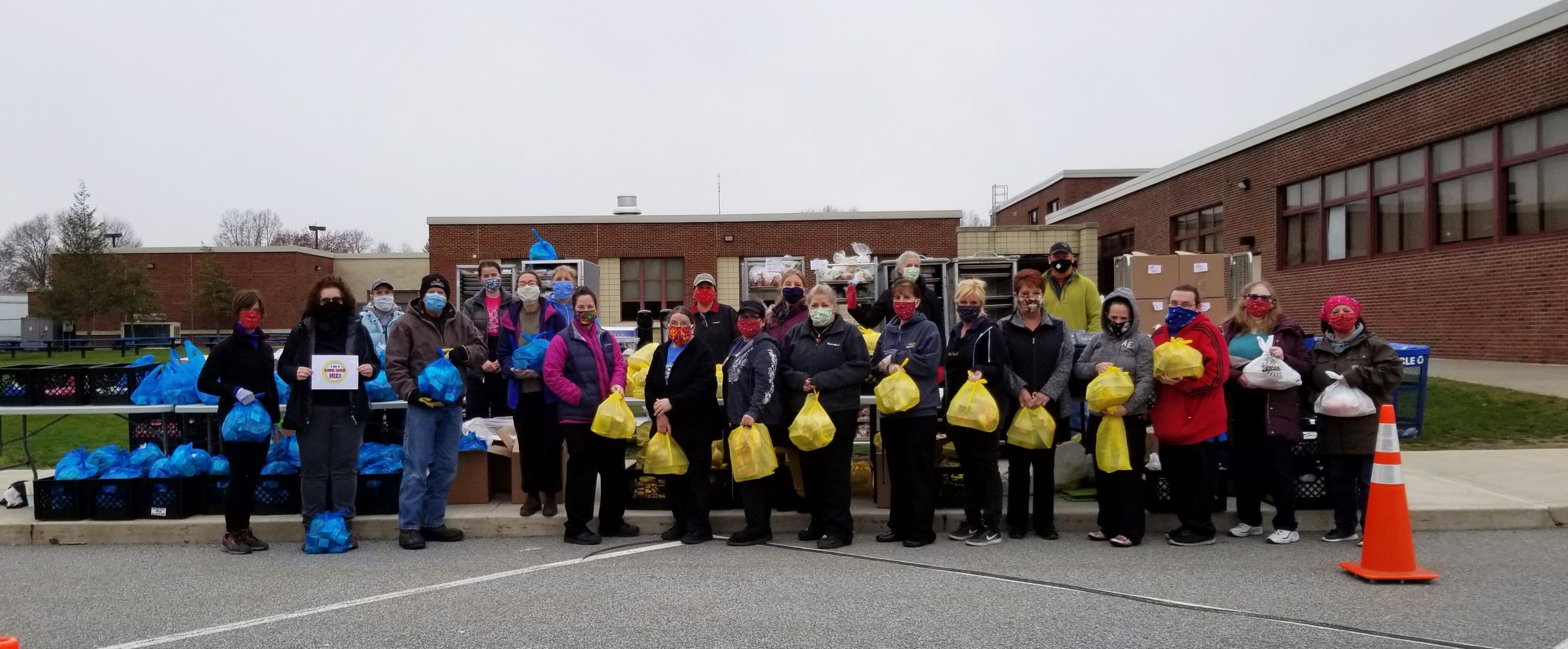 about 25 people, all wearing masks and holding plastic bags of food in front of a brick building