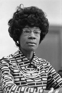 A woman with glasses, dark hair, wearing a blouse with geometric pattern. In black and white.