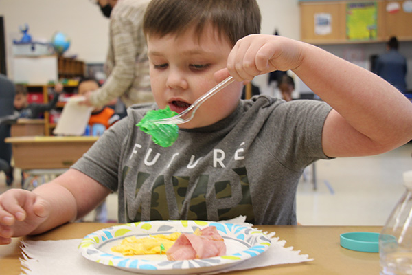 A young boy with short brown hair and wearing a gray shirt holds a fork up to his mouth with green scrambled eggs on it. A plate in front of him has ham and yellow eggs. His mouth is open.