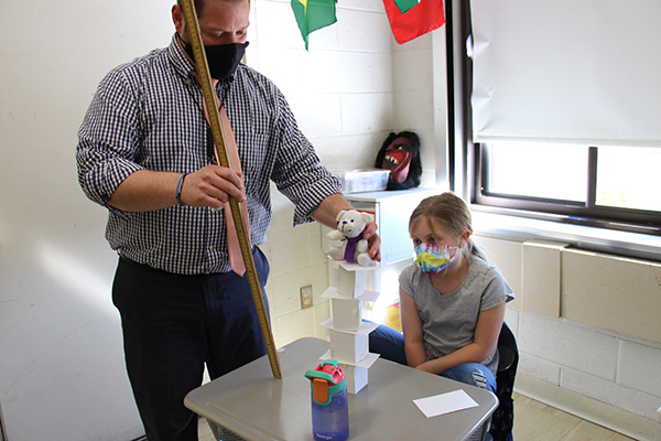 A man in a blue plaid shirt and light tie, wearing a dark face mask, places a small bear on top of a tower made of folded index cards. A little girl watches intently as she sits at her desk wearing a face mask and short sleeve shirt.