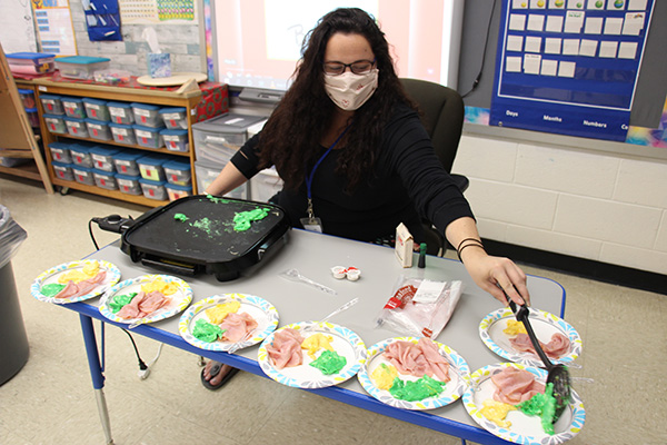 A woman with long dark hair, glasses and wearing a light colored face mask sits at a table with 7 plates all containing ham yellow and green scrambled eggs. She is using a spoon to put green eggs on one of the plates.