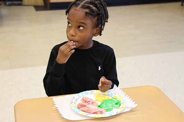 A young boy with short dark braids and wearing a black long-sleeve shirt tastes a piece of ham from the plate on his desk, which also contains green eggs.