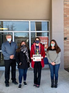 Four people - a man and three women - stand in front of a building. All are wearing masks and one is holding a certificate.