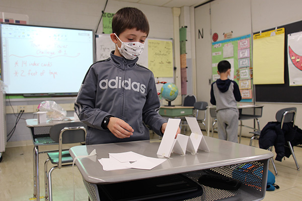 A boy wearing a gray sweatshirt and white mask stands at his desk that has several white index cards folded in triangles to make a structure.