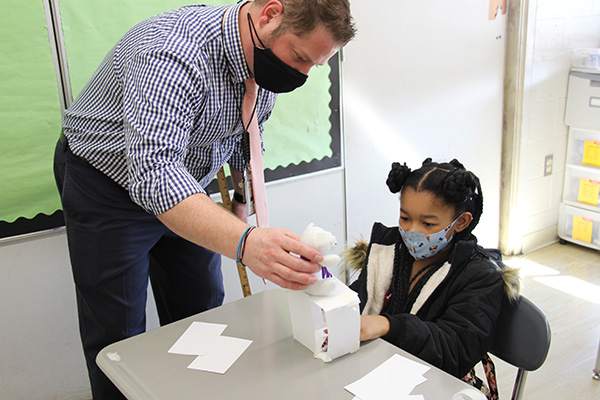 A man in a blue checked shirt an light tie wearing a dark mask leans over a desk with a small stuffed bear in his hand. He is placing it on a structure made of folded index cards. A girl with a blue mask and dark hair looks at it intently.