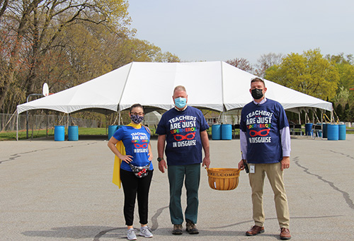 A woman and two men stand outside in front of a large white tent. They are all wearing shirts that say Teachers are just super heroes in disguise. The men are each holding a handle to a large wicker basket.