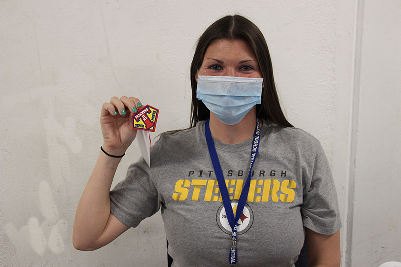 A woman with long dark hair and wearing a Pittsburgh Steelers t shirt and blue mask holds up her key chain that says Teaching is my super power.