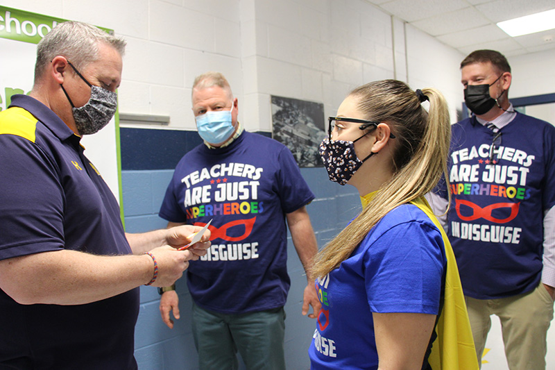 A woman with long hair in a ponytail, wearing a blue shirt and gold cape watches as a man in a blue shirt looks at the gift she just gave him. It's a keychain that says Teaching is my super power. Looking on are two other men wearing shirts that say Teachers are just super heroes in disguise. All are wearing masks.