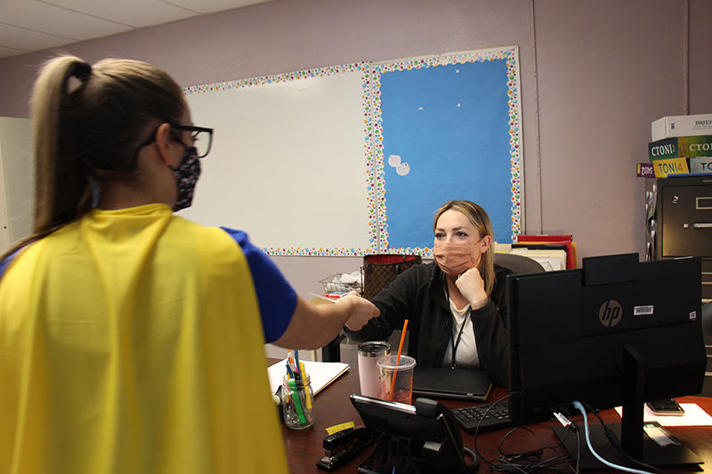 A woman sitting at a desk in a classroom reaches out her hand to take a key chain from a woman. The teacher at the desk is wearing a dark shirt and a facemask. The woman handing her the key chain is wearing a gold cape.