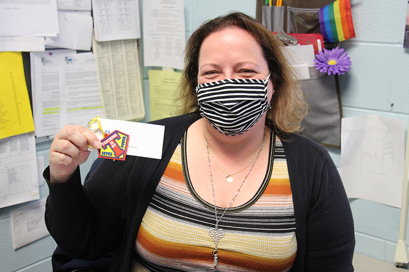 A woman broadly smiles behind her striped mask and holds up her key chain, given as a gift. The key chain says Teaching is my super power. She has short dark hair and is wearing a brown, beige and orange striped shirt.