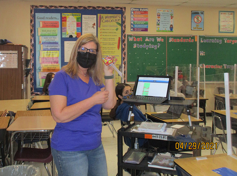 A woman with long blonde hair, wearing a black mask and purple shirt, holds up her key chain, she received as a gift. It says Teaching is my super power.