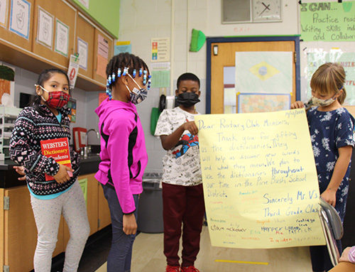 Two students, a boy and a girl, hold a large yellow poster with a thank you note for the Rotary. A girl in a pink jacket and beaded braids looks at the note and reads it. Another girl holds the red dictionary.