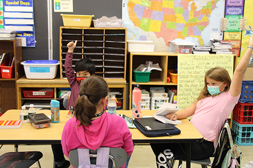 Three students - a girl with her backk to the camera, a girl on the right wearing a pink shirt and blue mask with her hand raised, and a boy with dark hair raising his hand with his thumb up. 