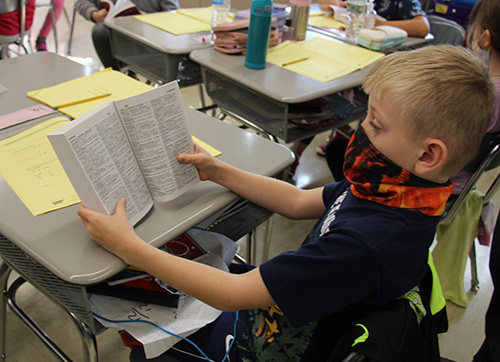 A boy wearing a dark shirt and a red and blue mask holds his new dictionary open.