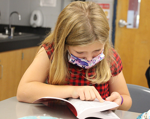 A girl with long blond hair, wearing a purple face mask and a red and black check shirt, has her dictionary open and uses her finger to point to a word.