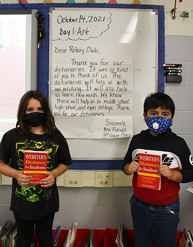 A large letter written on a large white poster paper is in the background. It thanks the Rotary for their gift of the dictionaries. In front are two students, a girl on the left with longer dark hair and a black shirt, wearing a black mask and holding a red dictionary. The boy on the right has short dark hair and is wearing a red white and black shirt, blue mask and is holding a red dictionary.