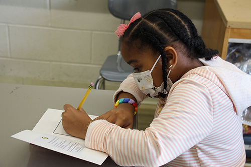 A girl with black braids, wearing a pink ribbon in her hair and a striped shirt, writes her name in her dictionary.