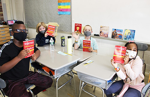 Four third grade students sit at their desks and hold up their red webster's dictionaries. All are wearing masks, one is waving.