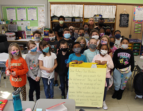 A class of 23 third-grade students stand together. Two in the front hold a large yellow poster with a note on it thanking the Rotary club for their gift of dictionaries.