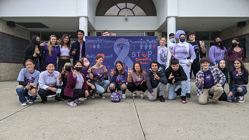 A group of 24 high school students squat and stand by a purple sign. They are all wearing some sort of purple and holding peace signs up. The sign says Domestic Violence Awareness Month. Real Love Doesn't Hurt and Stop Domestic Violence.