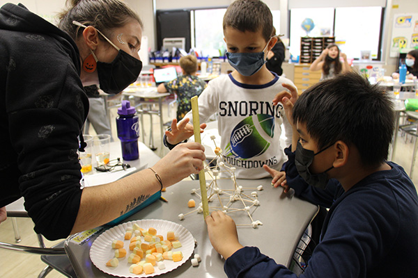 Two elementary students are at a table with mini marshmallows and toothpicks. There is a structure made with them. A woman wearing a black sweatshirt and mask, with pumpkin earrings holds a ruler up to the structure.