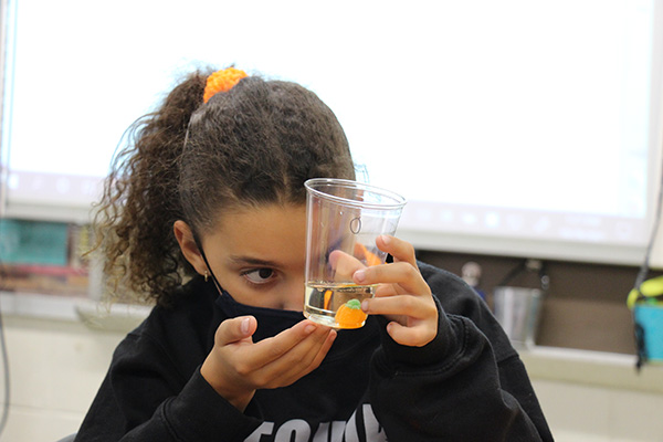 A girl with dark wavy hair pulled back in a ponytail holds a clear cup with a clear liquid and an orange pumpkin candy in it. She is holding it up and looing intensely at it. She is wearing a mask and a black sweatshirt.