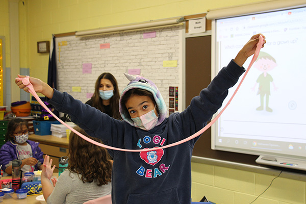 An elementary age student wearing a blue sweatshirt with the words Yogi Bear on it stretches pink homemade slime. She is wearing a light colored mask and there are other children and an adult in the background.