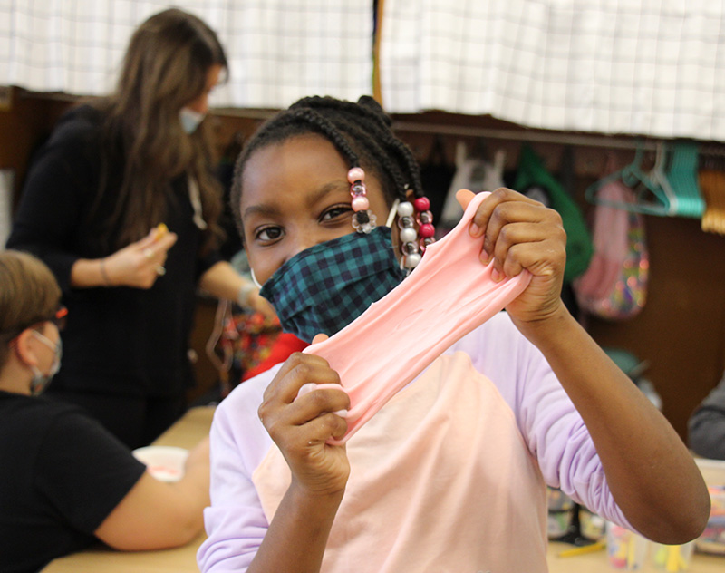 An elementary girl, with pink beads in her braids, stretches pink slime. She is wearing a green and blue check mask and a purple and peach shirt.