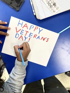 A blue desk with a white piece of paper on it. A student's hands are writing with a blue pencil. It says Happy Veterans Day!