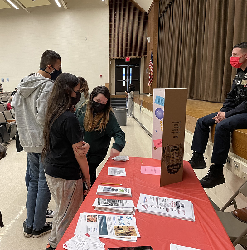 Students wearing masks gather near a table that has flyers on it explaining what clubs are available to them.