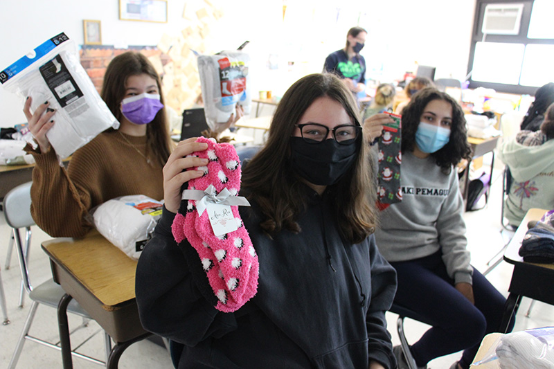 Three middle school girls hold up groups of socks they collected. All are wearing masks.