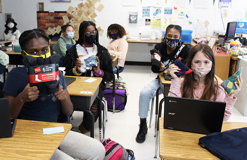 Four middle school girls hold up socks while sitting at their desks. 