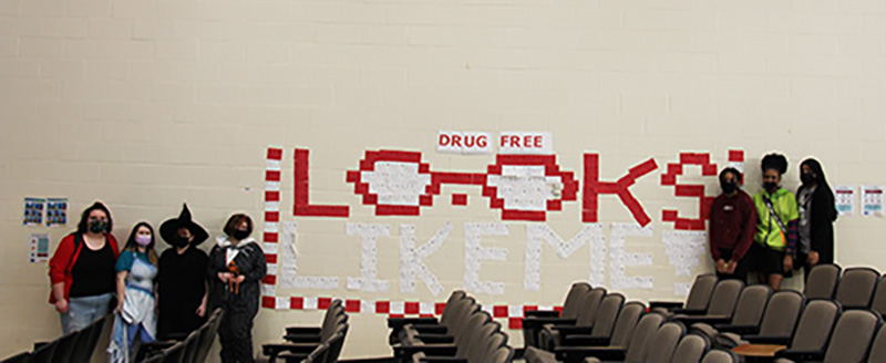 A white wall in the background with red postcards on it, in the shape of words that say Drug Free Looks like Me. There are four girls on the left and three on the right.