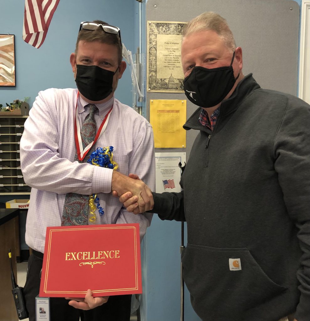 A man on the left, wearing a shirt and tie and a black mask, shakes hands with the man onthe right. The man on the left is holding a red certificate that says Excellence in gold. The man on the right is wearing a black jacket and mask.