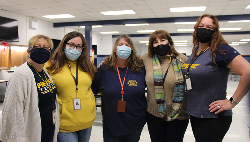 A group of five women stand together in a large room behind them. They have arms around each other's shoulders. They are wearing masks.