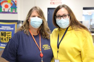 Two women, both with long brown hair, stand with each other. The one on the left is wearing a navy blue shirt with gold writing on it and a blue mask. She has an orange lanyard around her neck with an id badge on it. The woman on the right is wearing glasses, a gold sweatshirt, blue lanyard with id badge. They both have light blue masks on.