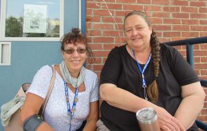 Two women smile. They are outside of a brick building. The woman on the left is wearing a light colored shirt and her hair is pulled up into a bun. She has a lanyard around her neck. The woman on the right is wearing a black shirt. Her hair has a long braid over her shoulder. She too has a lanyard on and is holding a cup.