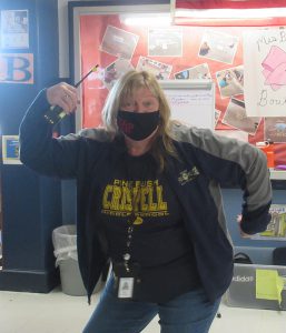 A woman with shoulder-length blonde hair, wearing a blue and gray jacket and a blue and gold shirt that says Crispell, strikes a pose with her arms up showing muscles.