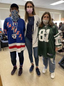 Three women stand together, all wearing masks. They all have sports jerseys on, one a Rangers jersey, one a baseball jersey and one a jets jersey.