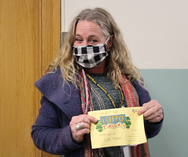 A woman with long blonde hair wearing a black and white checked mask. She is holding a yellow piece of paper that says We hit the Lotto with your on our team.