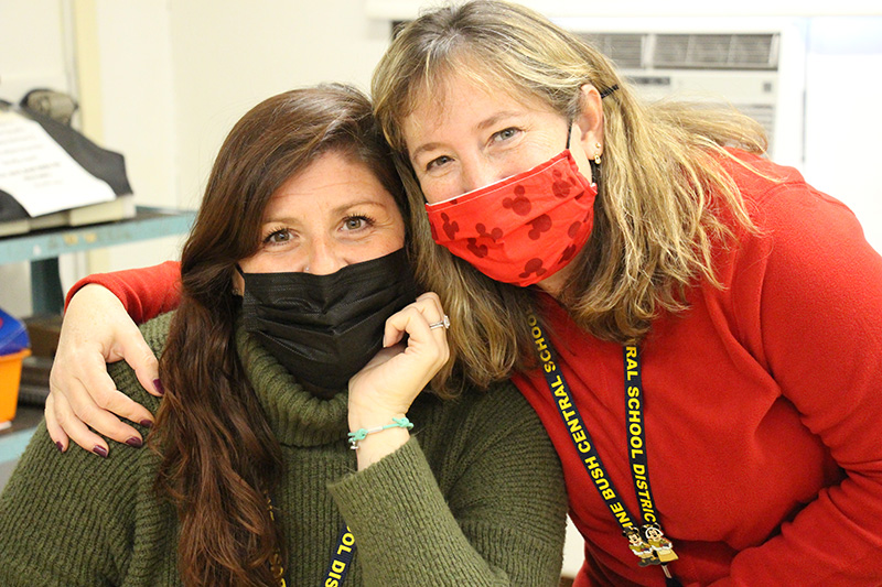 Two women  stand with each other, the one on the right with her arm around the other. They are both wearing masks. The woman on the left has long dark hair and is wearing a green sweater. The woman on the right is wearing an orange sweater and has shoulder-length blonde hair.