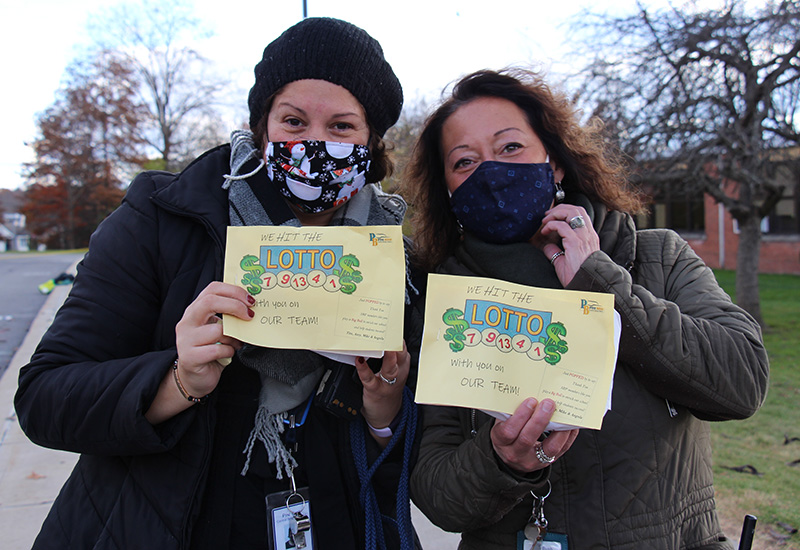 Two women lean in to each other. They are outside and wearing winter jackets, holding up yellow papers that say We hit the Lotto with you on our team.